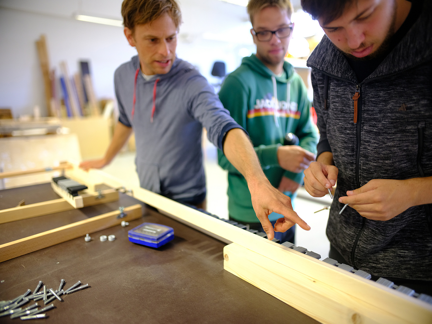 Fachanleiter zeigt zwei Beschäftigten etwas. Sie bauen ein Rahmen aus Holz
