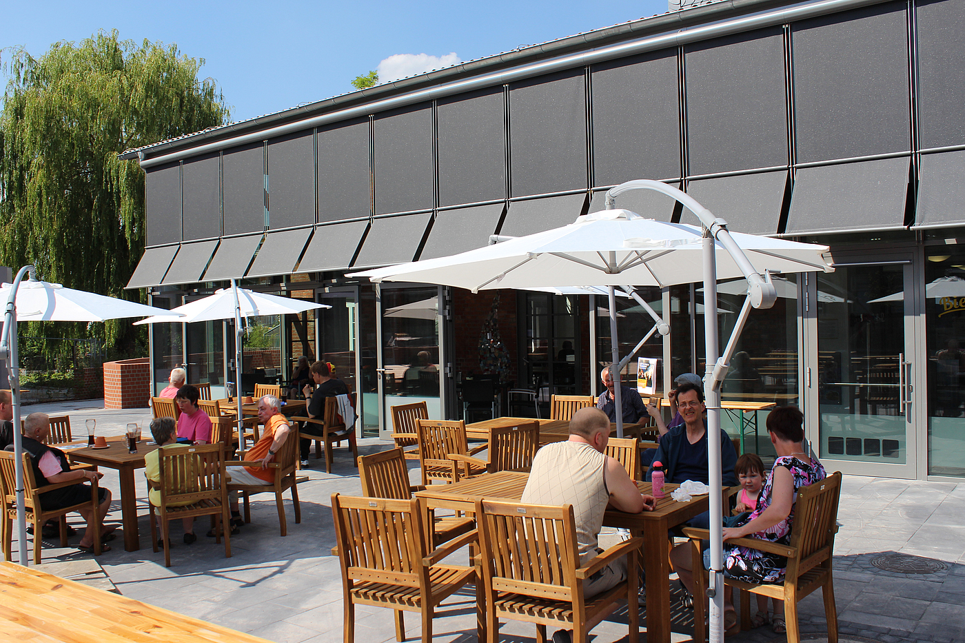 Gebäude des "Biermanskis" von Außen. Sitzgruppen mit weißen Sonnenschirmen und Menschen, die Essen und das Wetter genießen