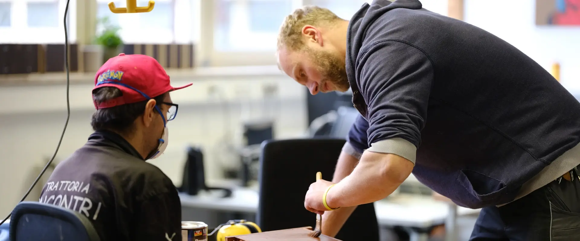 Fachanleiter zeigt einem Teilnehmer mit roter Cappy und schwarzer Kleidung wie etwas gestrichen werden kann